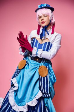 Mavi şapkalı, parlak elbiseli, pembe arka planda kameraya bakan çekici bir bayan Cosplayer.