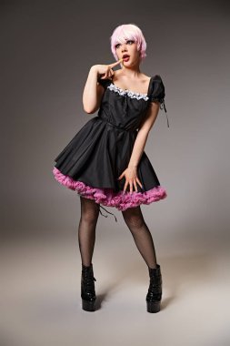 Siyah elbiseli, pembe saçlı, gri arka planda poz veren ve başka yöne bakan seksi kadın bir Cosplayer.