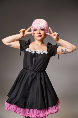 cheerful alluring female cosplayer in black dress showing peace gestures and smiling at camera clipart