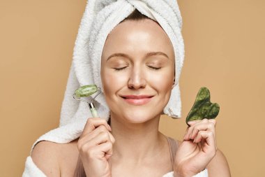 A serene woman with a towel on her head holds a face roller and a, showcasing her natural beauty routine. clipart
