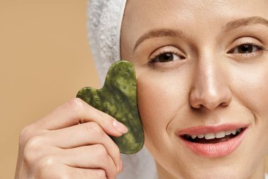 Doğal güzellikte bir kadın, bu canlandırıcı ve eğlenceli sahnede kafasında bir havlu dururken neşeyle gua sha tutar..