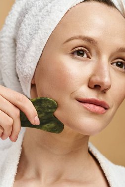 A woman, showcasing natural beauty, holds a gua sha while a towel rests on her head. clipart