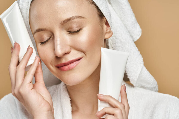An attractive woman with a towel wrapped around her head holding a tube of cream, focused on her skincare routine.