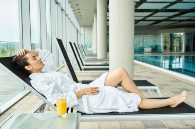 A young, beautiful brunette woman is lounging on a lounge chair next to an indoor swimming pool, basking in relaxation. clipart