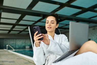 Genç ve güzel bir esmer kadın, kapalı bir spa havuzunun yanında bornozla rahatlar ve cep telefonuna odaklanır..