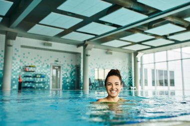 A young brunette woman in a swimsuit smiles while submerged in an indoor spa swimming pool. clipart