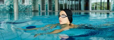 Mayolu ve yüzme şapkalı bir kadın gözlüklü bir havuzda yüzüyor ve suda dalgalanmalar yaratıyor..