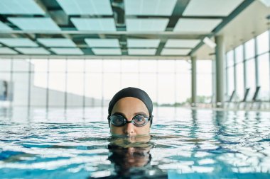 Genç kadın gözlük ve yüzme şapkasıyla havuzda süzülüyor, akışkan hareket ve sükuneti somutlaştırıyor..