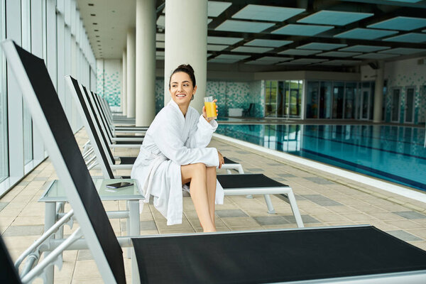 A young brunette woman sits on a lounge chair by an indoor pool, holding a drink in her hand.