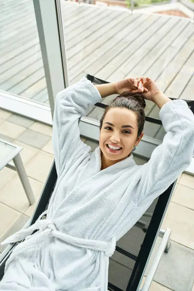 stock image A young, beautiful brunette woman in an indoor spa, wearing a bathrobe, sitting gracefully on a lounge chair, by the swimming pool.