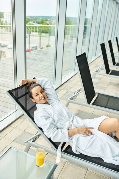 A young, beautiful brunette woman reclining on a lounge chair in a tranquil indoor spa setting near a swimming pool.