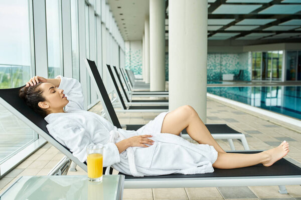 A young, beautiful brunette woman is lounging on a lounge chair next to an indoor swimming pool, basking in relaxation.