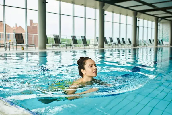 stock image A young brunette woman in a swimsuit gracefully swimming in a large indoor swimming pool.