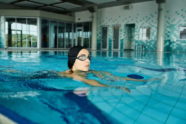 一名身穿泳衣 头戴泳帽的年轻女子潜入室内温泉池 戴着护目镜 以获得清晰的水下视力 — 图库照片