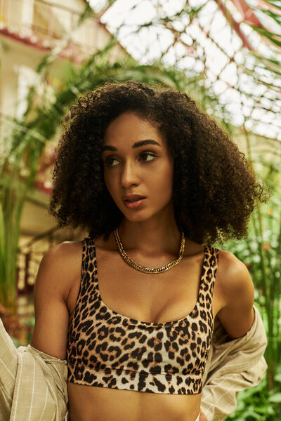 Fashionable african american woman with curly hair in animal print look in tropical greenhouse