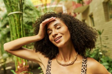 happy black woman in animal print top posing with hand near curly hair in atrium with green plants clipart
