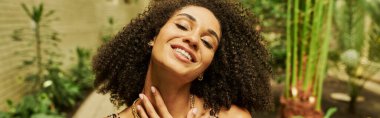 happy black woman in animal print top posing with hand near neck in atrium with green plants, banner clipart