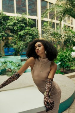 Afro-Amerikan elbiseli ve eldivenli çekici kadın şehir bahçesindeki bitkiler arasında poz veriyor.
