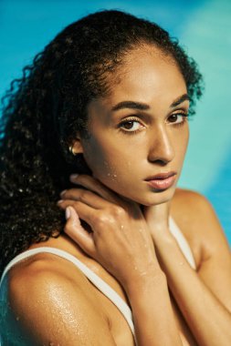 Genç siyah, kıvırcık saçlı bir kadın serin mavi havuz suyunda dikilip kameraya bakıyor.