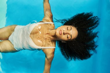 Mavi havuz suyunda sırt üstü yüzen rahat Afro-Amerikan kadın manzarası. Huzur.