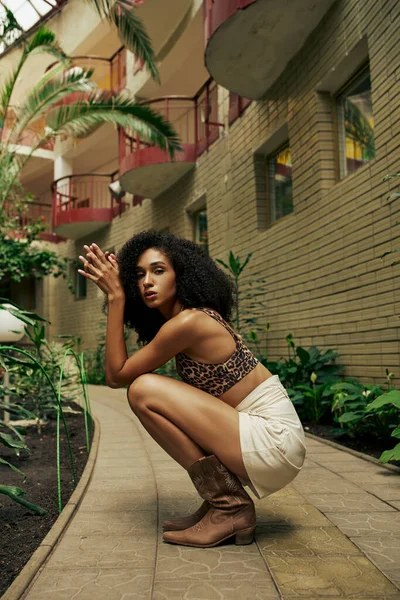 シックな服装の美しいアフリカ系アメリカ人カーリー女性が緑の植物と建物の近くにポーズを打つ — ストック写真
