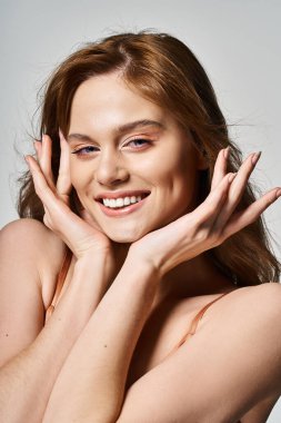 Cheerful smiling woman with trendy makeup, looking at camera, hands near face on grey background clipart