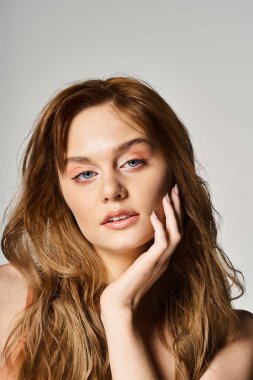 Mavi gözlü, yanağına dokunup gri arka planda poz veren seksi, hayalperest kadının portresi.