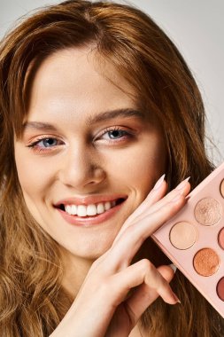Portrait of a beautiful smiling woman holding nude eyeshadow palette near face and looking at camera clipart