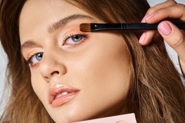 Gri arka plana makyaj ve fırçayla göz farı süren güzel bir kadın portresi.