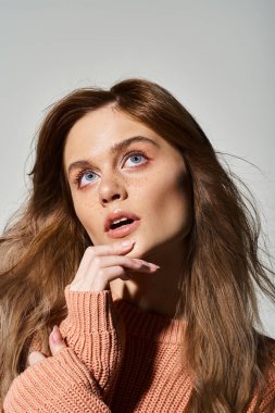 Düşünceli bir kadın çıplak şeftali makyajıyla yukarı bakıyor, kazak giyiyor, çenesine dokunuyor, gri arka planı var.