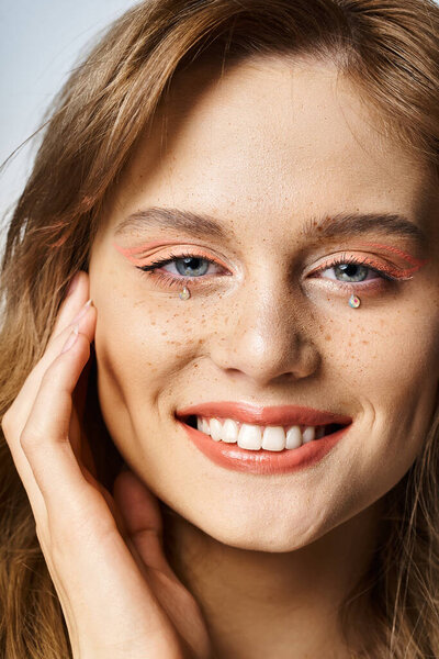Closeup beauty portrait of smiling girl with tear face jewels, peach makeup and freckles on grey