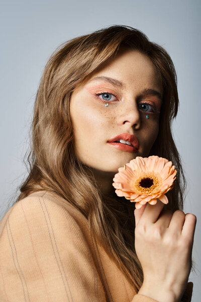 Красивая женщина с драгоценностями на лице в бежевой куртке, держа маргаритку персикового цвета на сером фоне