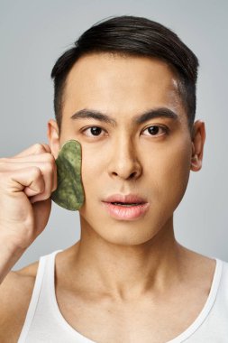 Asian man using green gua sha on his face in a grey studio setting. clipart