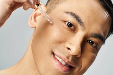 Yakışıklı Asyalı adam gri stüdyoda nazikçe deri pipetini serumla tutuyor..