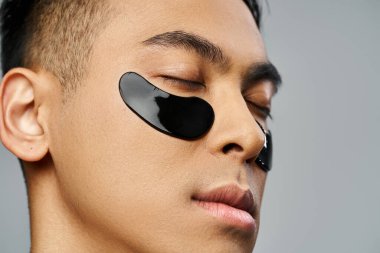 Handsome Asian man in a beauty routine, wearing a black eye patch in a grey studio setting. clipart