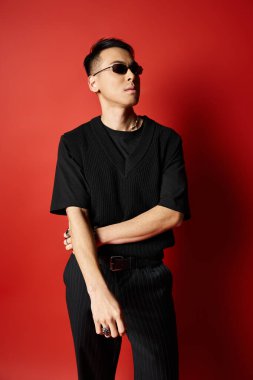 A stylish and handsome Asian man dressed in a black shirt and black pants poses against a bold red background in a studio. clipart