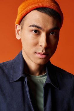 Şık mavi gömlekli ve turuncu şapkalı yakışıklı bir Asyalı stüdyoda turuncu arka planda duruyor..