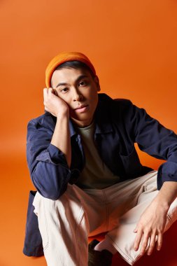 Yakışıklı bir Asyalı, şık giyinmiş, bir taburede oturuyor ve eli başının üstünde düşünceli bir şekilde duruyor..
