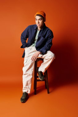 Zarif giyinmiş yakışıklı bir Asyalı ahşap bir sandalyenin üzerinde turuncu bir arka planda oturuyor..