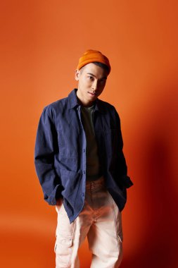 Mavi gömlekli ve turuncu şapkalı yakışıklı Asyalı bir adam stüdyo ortamında turuncu bir arka plana karşı şık bir şekilde poz veriyor..