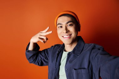 Stylish Asian man in a blue jacket and orange hat poses confidently against an orange background in a studio. clipart