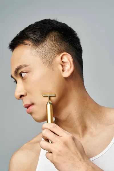 一个英俊的亚洲男人在灰色的工作室 拿着一把金色的剃须刀 望着别处 — 图库照片