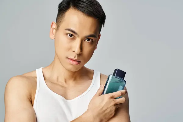 タンクの上のハンサムなアジアの男は繊細に灰色のスタジオの設定で香水の瓶を保持しています — ストック写真