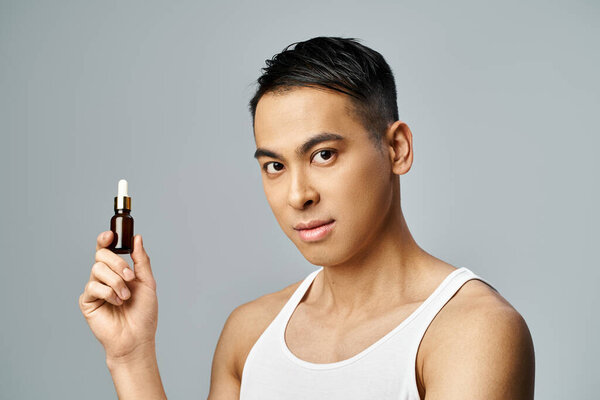 A handsome Asian man holds a bottle of oil in a grey studio, showcasing his skincare routine with precision and style.