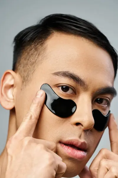 stock image A handsome Asian man in a grey studio, using black eye patches as part of his beauty and skincare routine.