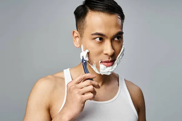 A portrait of a handsome Asian man with shaving foam on his face, carefully shaving with a razor in a grey studio.