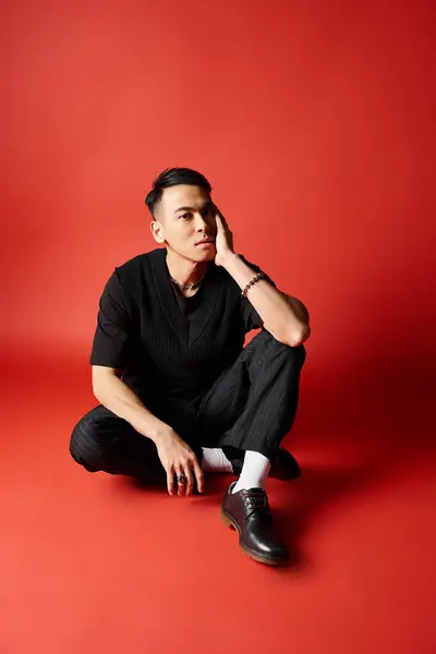 一位身穿黑色服装的时髦亚洲男子优雅地坐在地上 衬托着充满活力的红色背景 — 图库照片
