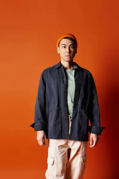 stock image A handsome Asian man stands confidently in stylish attire against a vibrant orange wall in a studio setting.