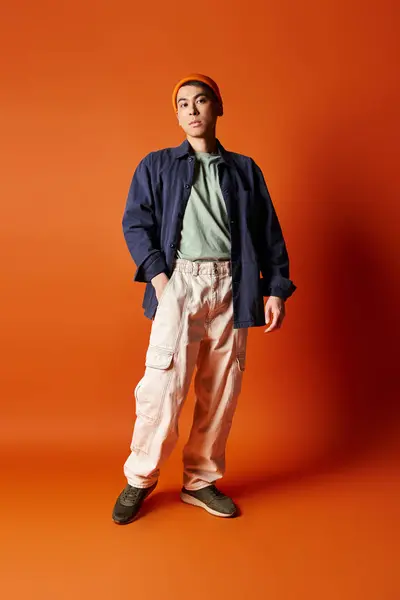 ハンサムなアジア人男性はスタジオの設定で活気に満ちたオレンジ色の背景に対してスタイリッシュな服装で自信を持って立っています — ストック写真