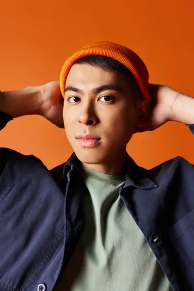 stock image A handsome Asian man in a blue jacket and orange hat, exuding style and elegance against a vibrant orange background.